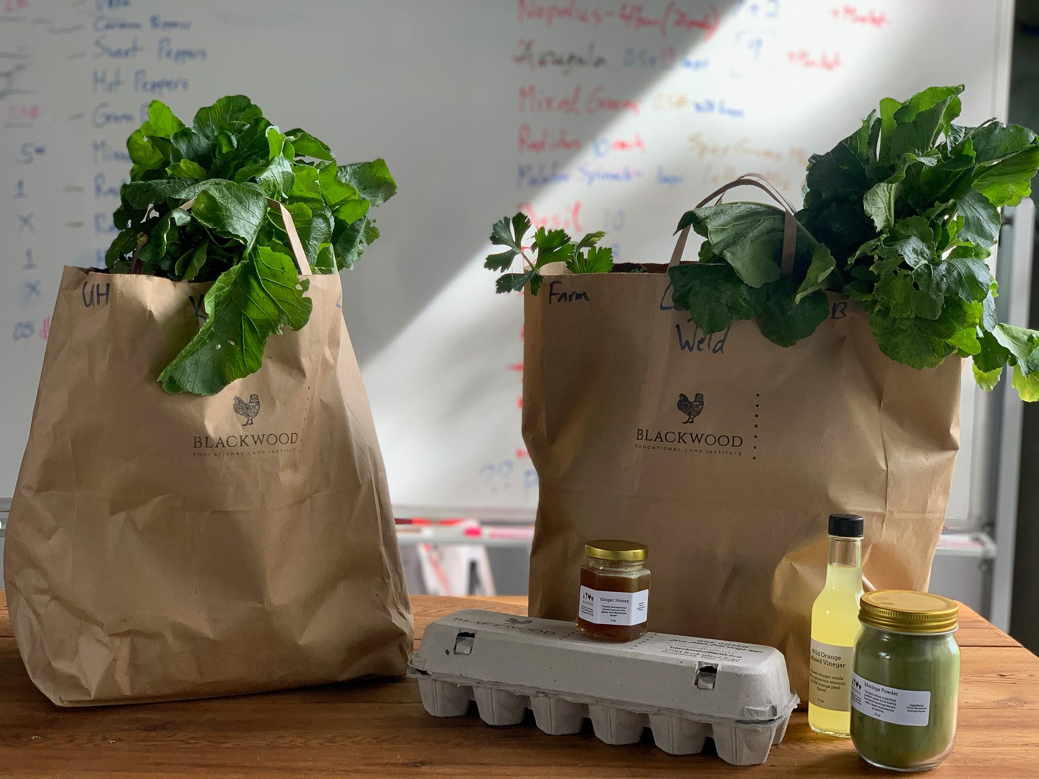 Two paper farm-share bags of leafy greens beside a carton of eggs, honey and oil
