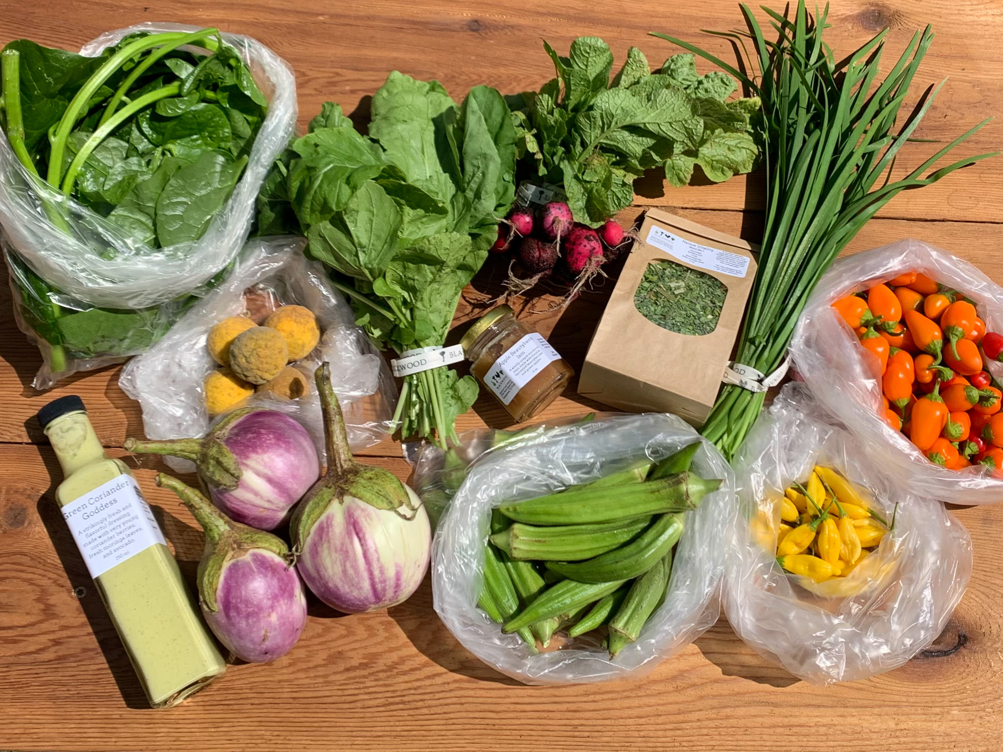 Overhead flat-lay of a harvest or farm-share haul arranged on a light wood table: a bag of spinach, a bunch of dark leafy greens, radishes with
