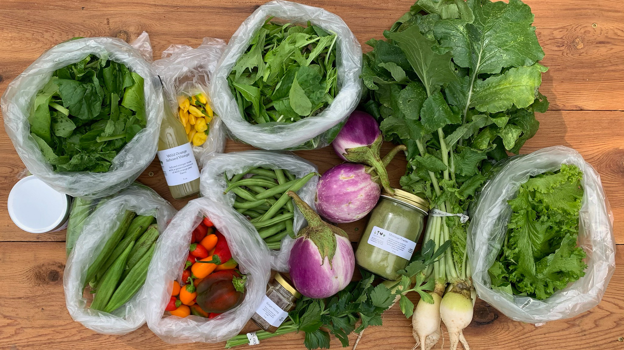 Overhead flat-lay of a harvest or CSA share on a wood table: bagged bok choy, bagged arugula, a bunch of large mustard or turnip greens with a