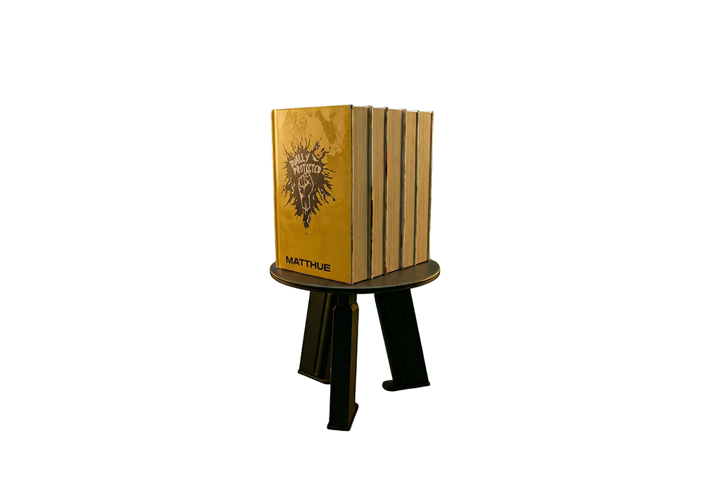 Five thick books standing upright on a small black stool with three legs. The front book has a yellow cover with an illustration of a raised fist and the words 'Fully Protected' above it and 'Matthue' at the bottom.