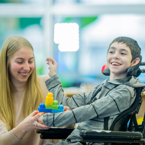 Smiling boy in wheelchair building with colorful blocks assisted by a cheerful woman.