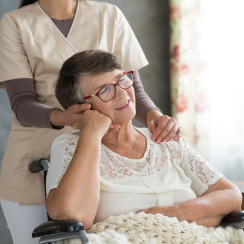 Smiling elderly woman in wheelchair being comforted by a caregiver in beige uniform.