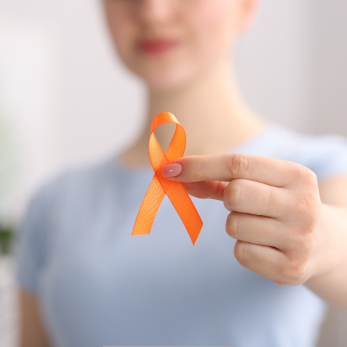 Person holding an orange awareness ribbon between thumb and index finger, blurred face in background.