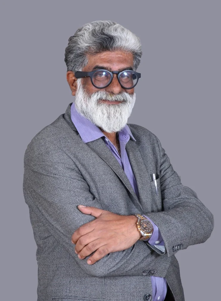Older man with white beard and hair wearing glasses, a gray blazer, and a purple shirt, arms crossed.