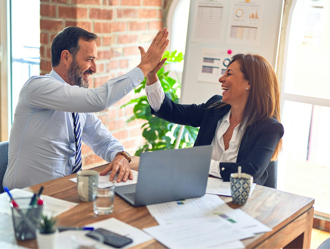 Two people sitting at a desk with papers all over it high-fiving