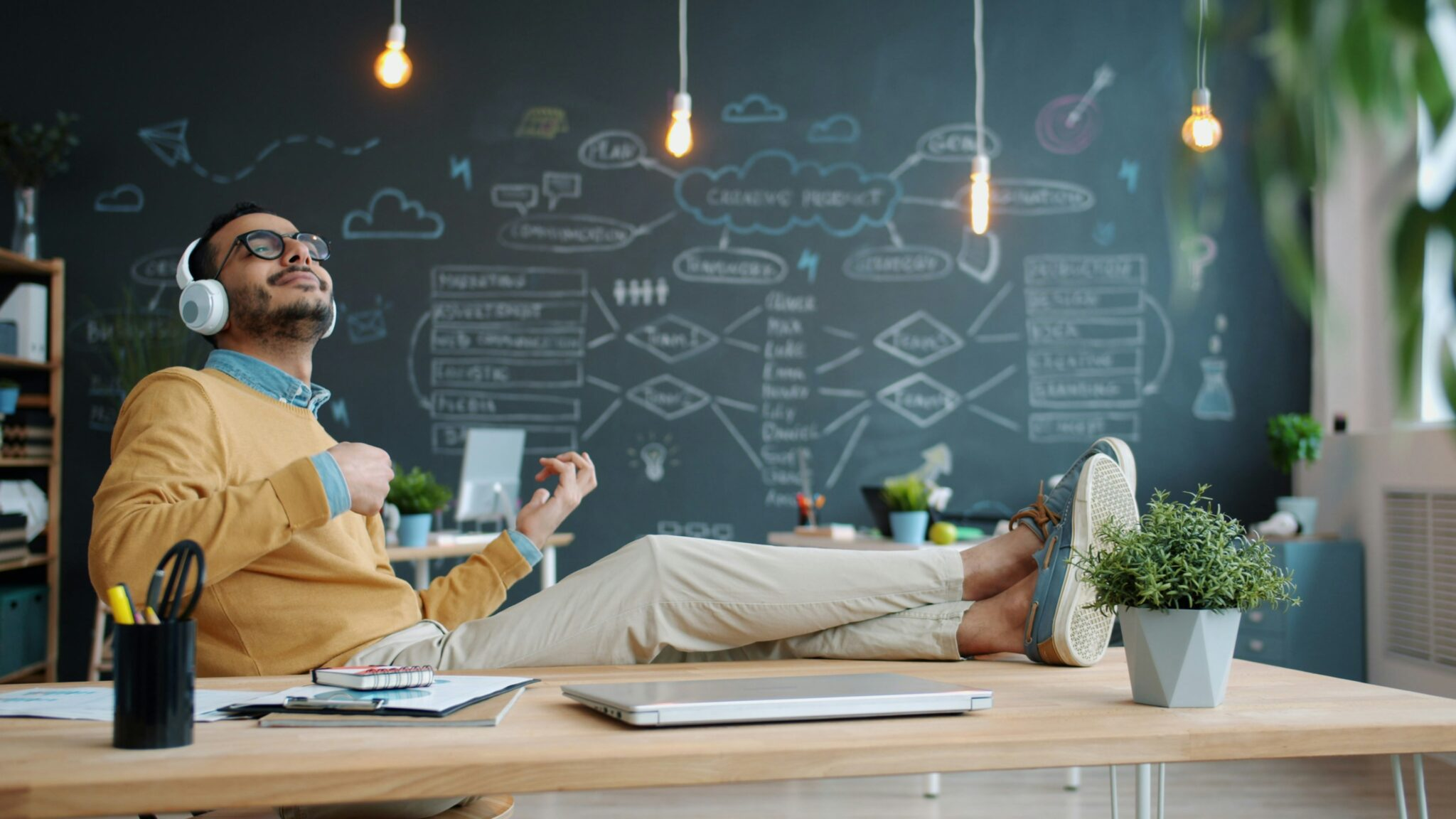 A man sitting at a desk with his legs up on it, looking up with his eyes closed. The man is also holding a cup of coffee