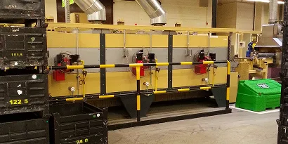Industriemaschine in gelber Farbe mit schwarzen Containern und grünem Abfallbehälter in Lagerhalle.