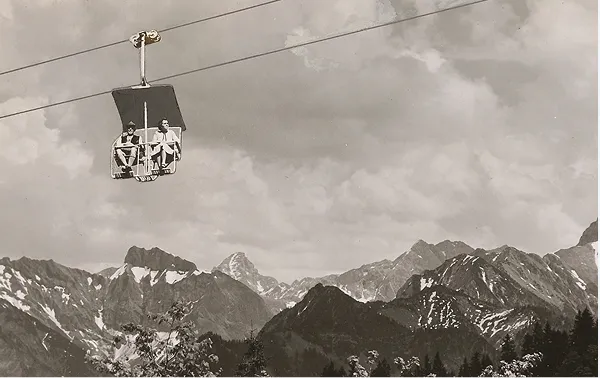 Zwei Personen sitzen in einer Gondel über schneebedeckten Bergen bei bewölktem Himmel.