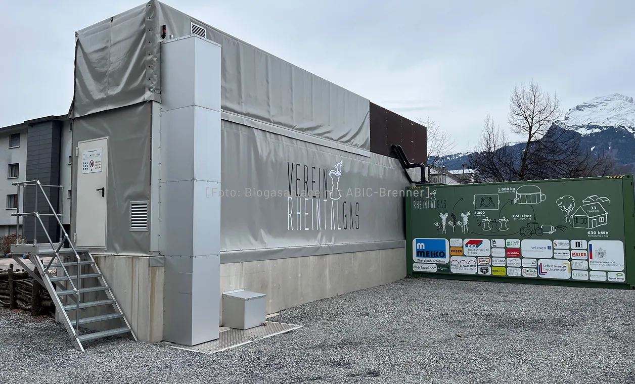 Biogasanlage mit grauer Plane und grüner Tafel zeigt Logos und Diagramme vor Bergkulisse.