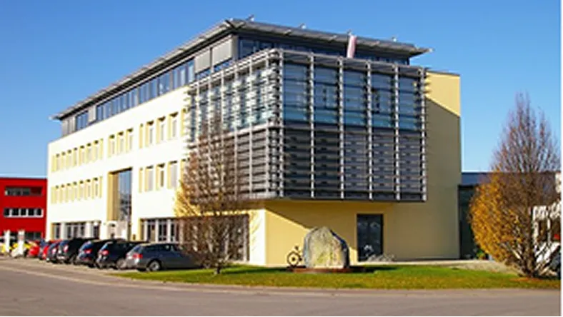 Modern three-story office building with yellow facade, glass windows, cars parked outside, and leafless trees.
