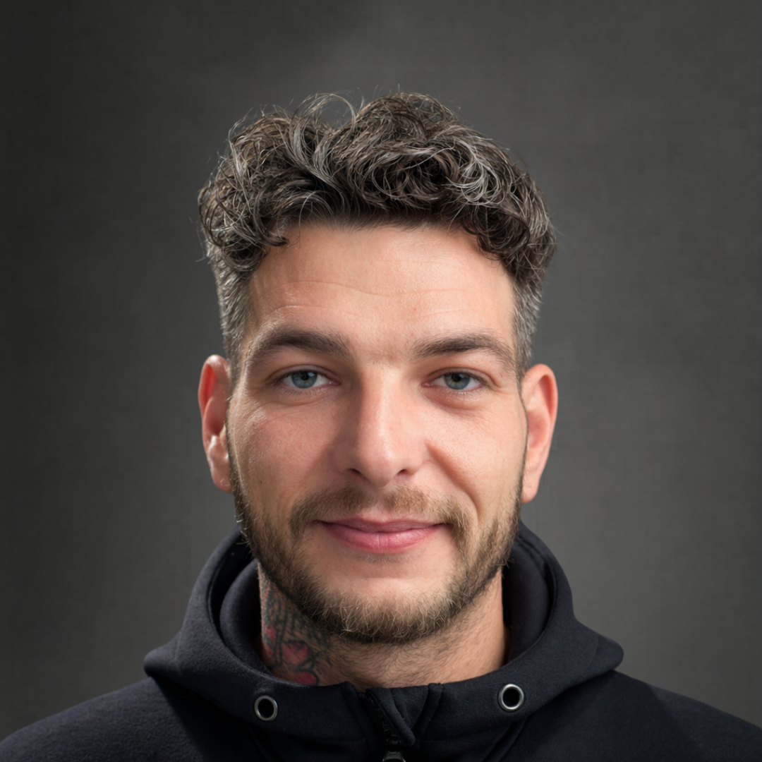 Portrait eines lächelnden Mannes mit lockigem Haar, Bart und schwarzer Kapuzenjacke vor grauem Hintergrund.