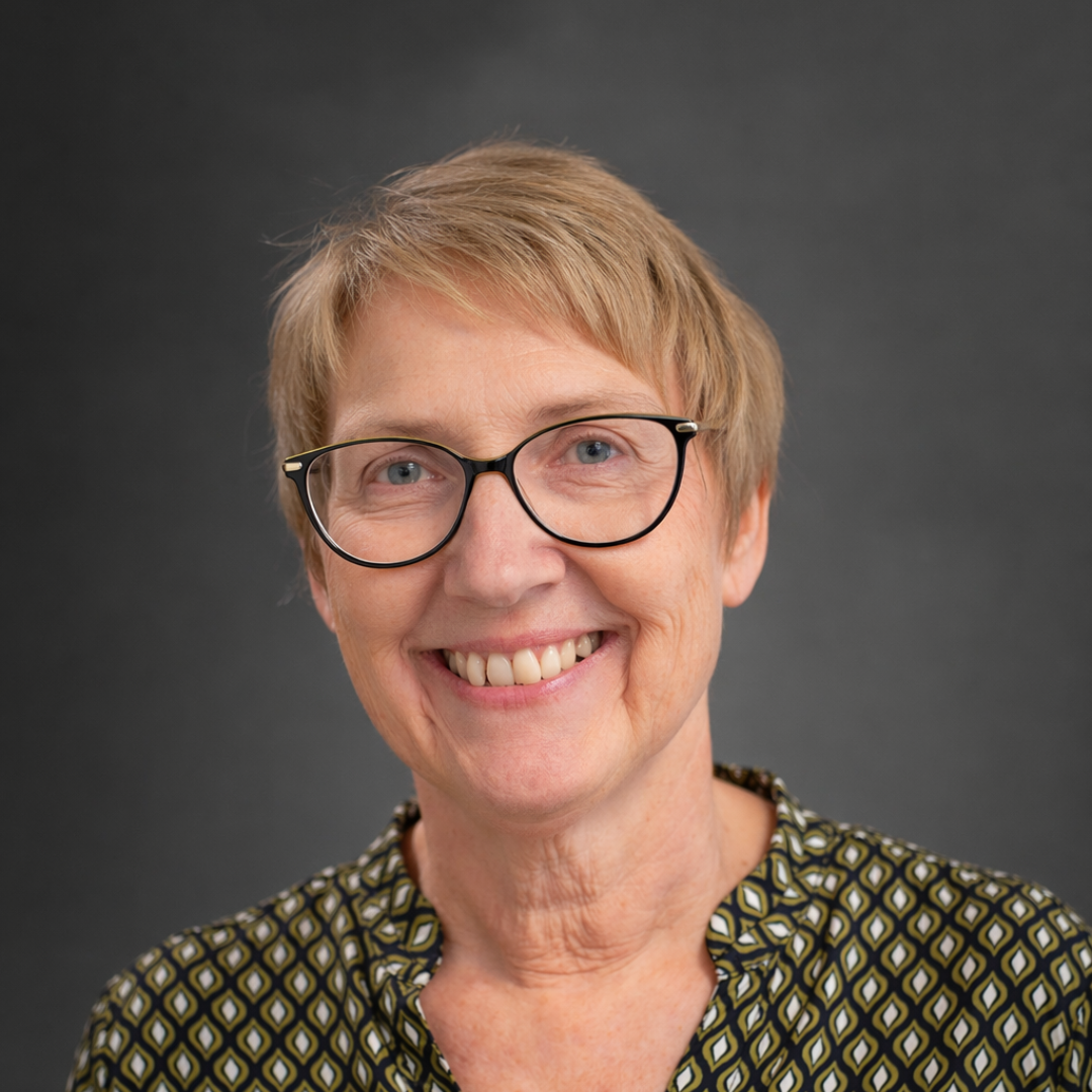 Ältere Frau mit kurzen blonden Haaren, Brille und gemustertem Oberteil lächelt vor grauem Hintergrund.