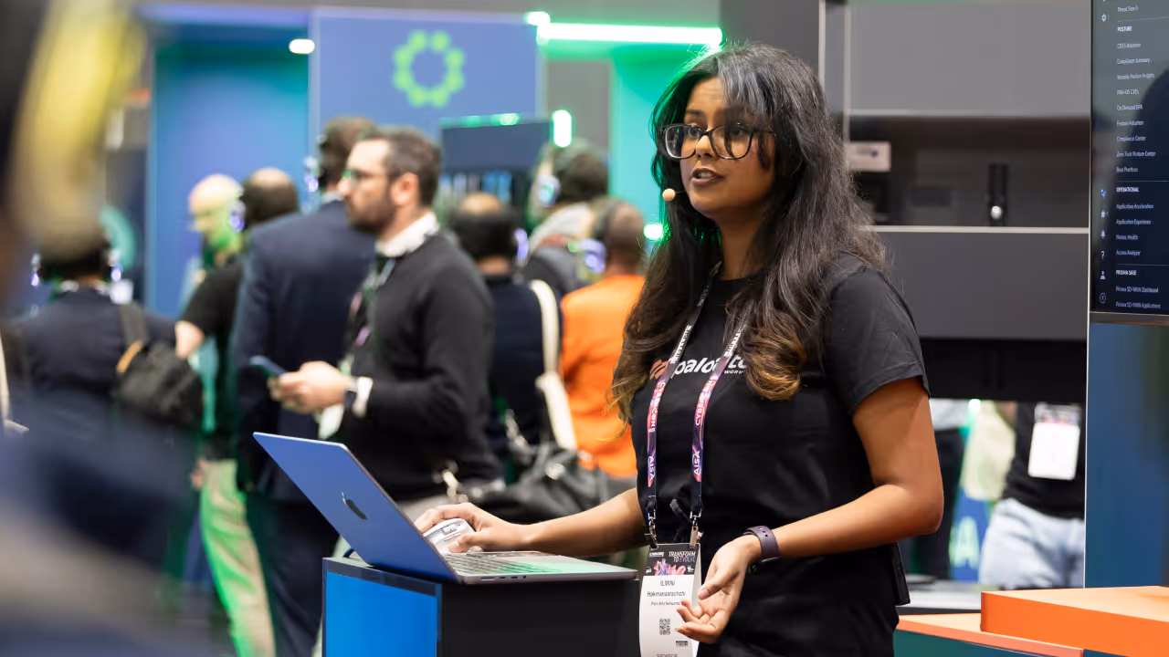 Woman with glasses speaking at a tech event, using a laptop on a podium with attendees behind her.