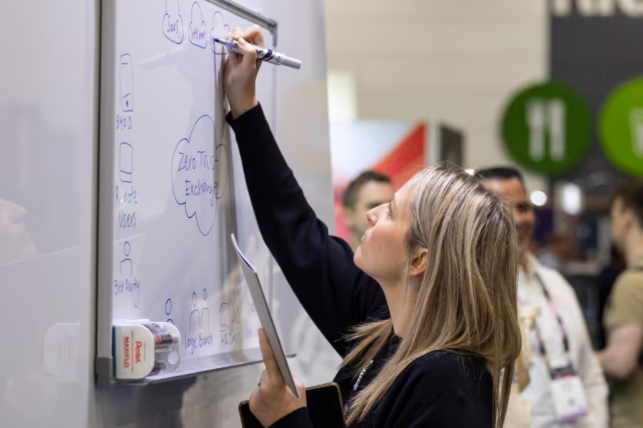 Woman writing on whiteboard with marker while holding a tablet, with people blurred in the background.