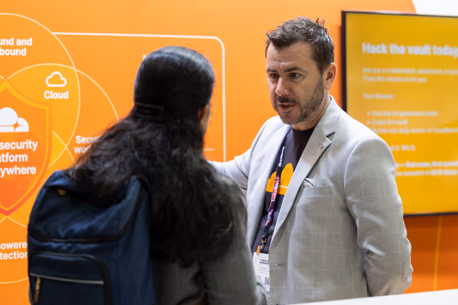 Man in light suit talking to a person with long hair and a backpack in front of an orange tech display.