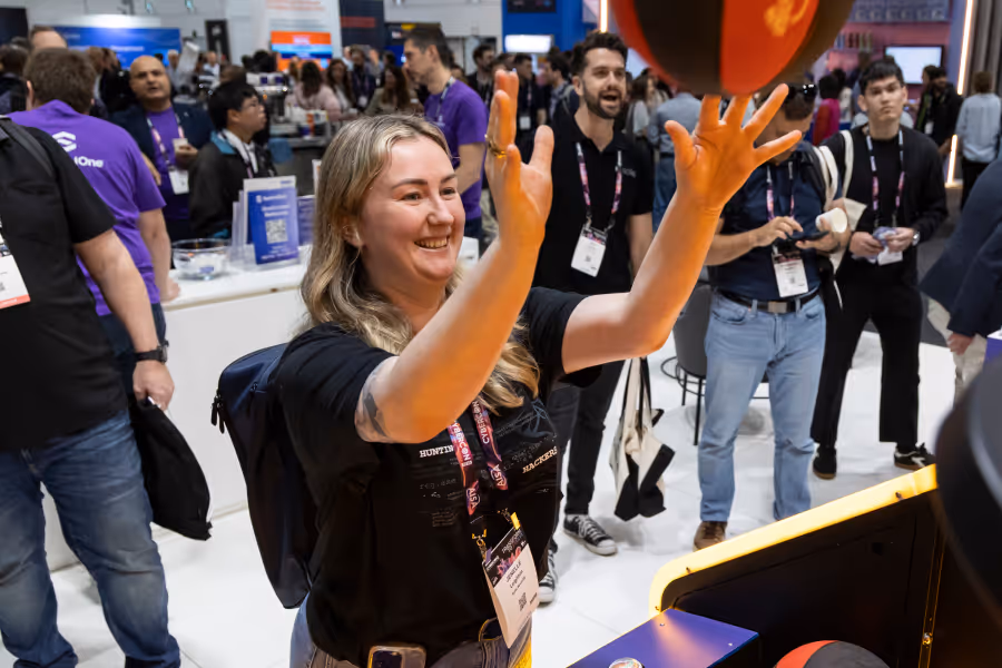 Smiling woman plays arcade basketball game at an indoor event with people watching nearby.