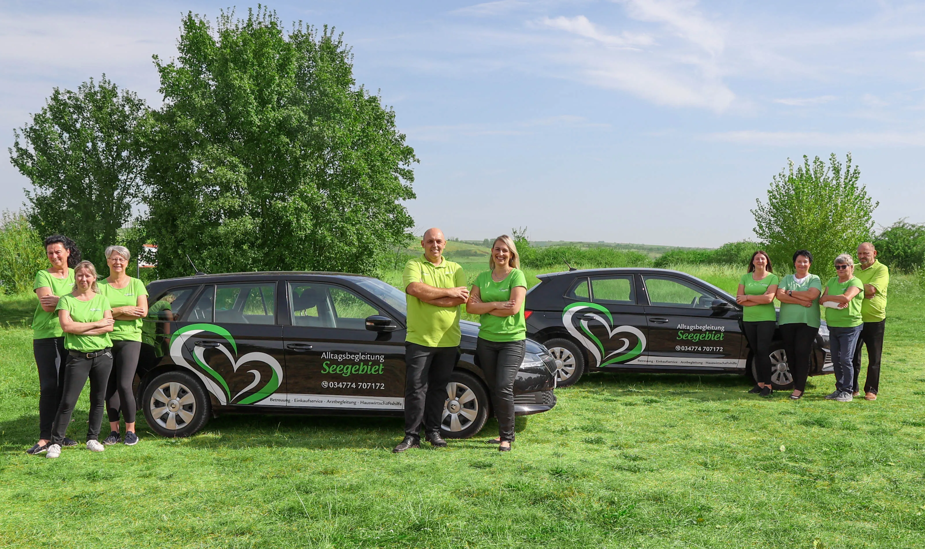 Group of ten people wearing green shirts standing with two black cars branded with 'Alltagsbegleitung Seegebiet' on a grassy field with trees.