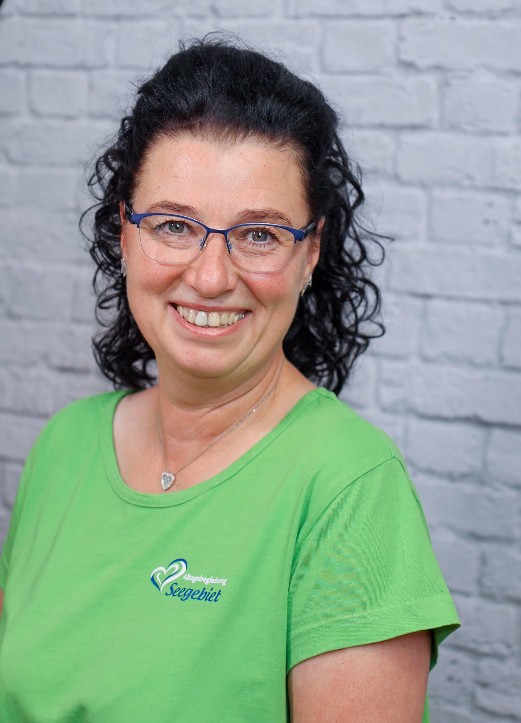 Smiling woman with glasses and curly dark hair wearing a bright green shirt with a heart logo.