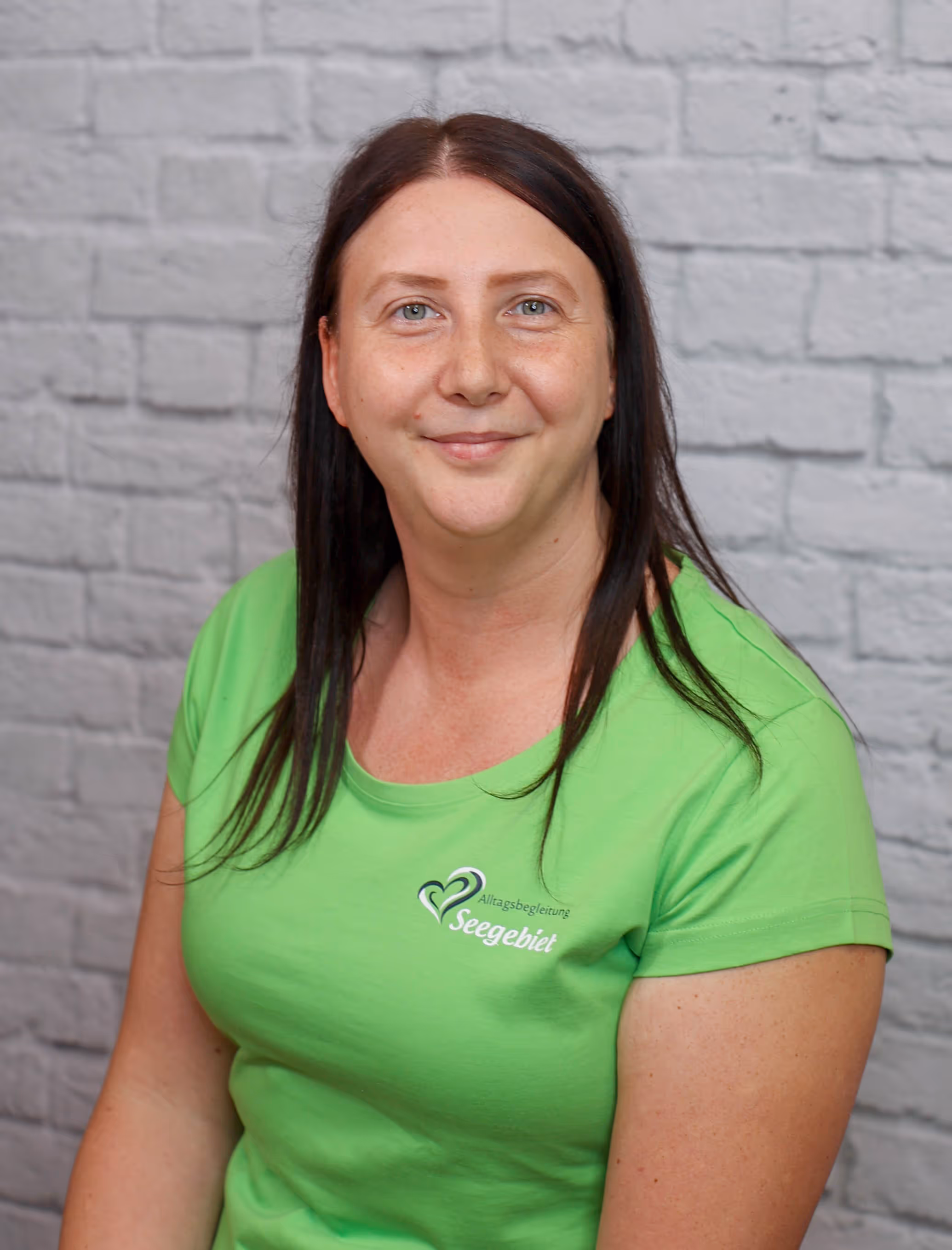Smiling woman with long dark hair wearing a bright green Seengebiet shirt against a gray brick wall.