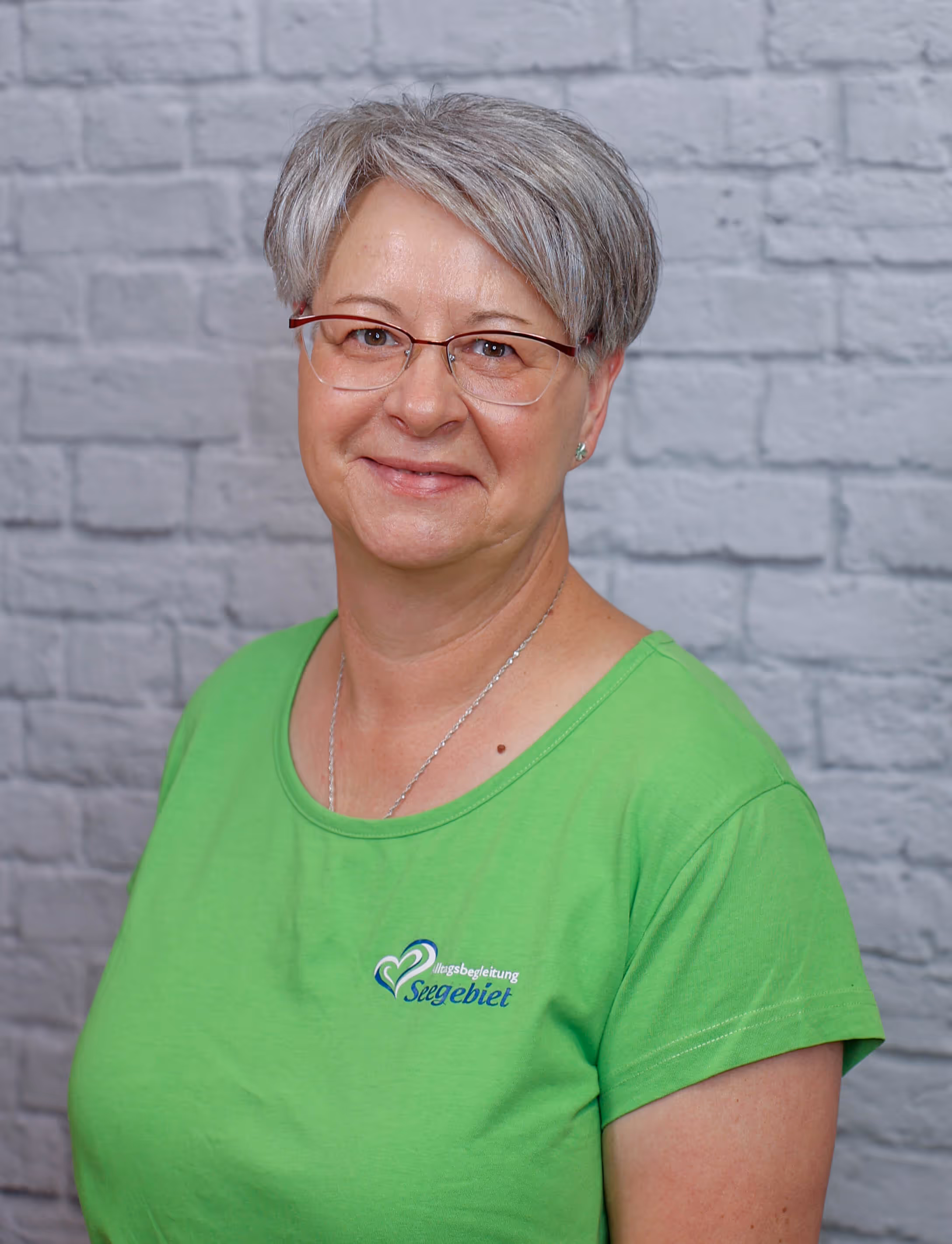 Smiling woman with short gray hair and glasses wearing a green shirt with a logo, standing against a gray brick wall.