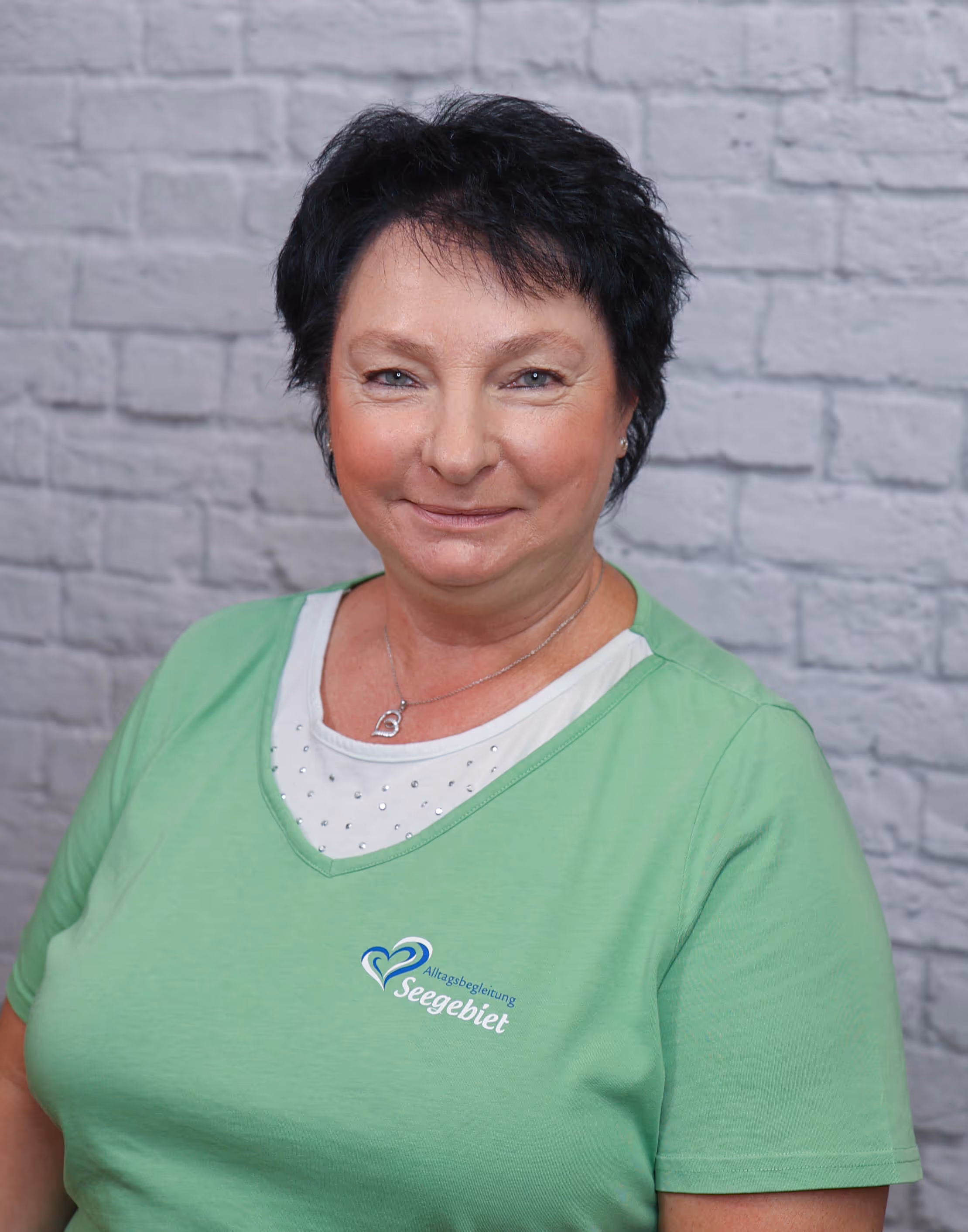 Smiling woman with short dark hair wearing a green shirt with a logo against a gray brick wall.
