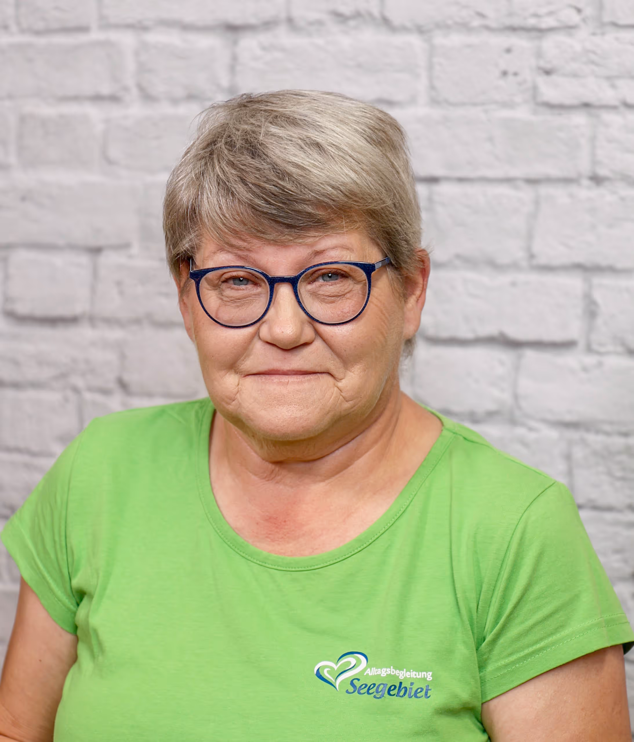 Woman with short gray hair and glasses wearing a green shirt with 'Seengebiet' logo against a brick wall.