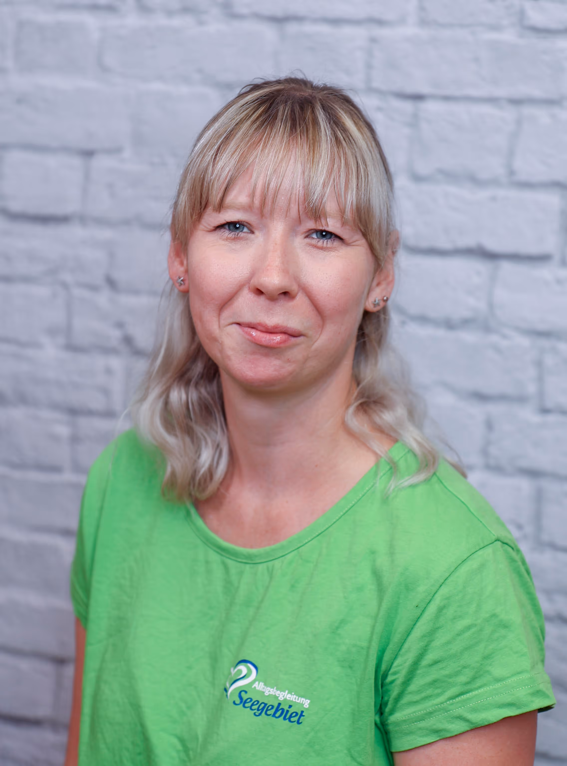 Woman with blonde hair and bangs wearing a bright green Seengebiet t-shirt against a gray brick wall background.