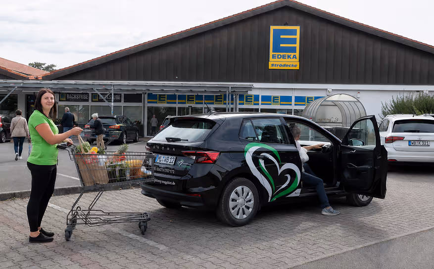 Woman with shopping cart full of groceries near a parked black car with heart logo in front of EDEKA store.