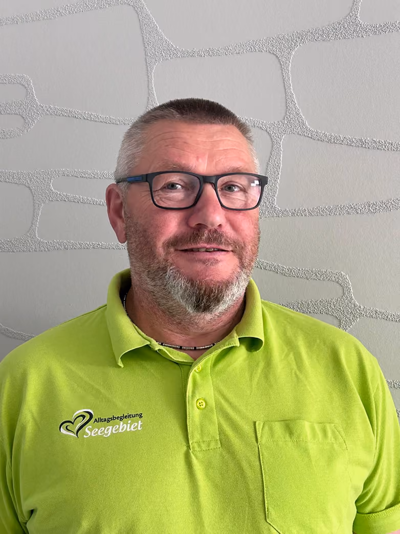 Man with glasses and short beard wearing a bright green polo shirt with Seegebiet logo on light textured wall.