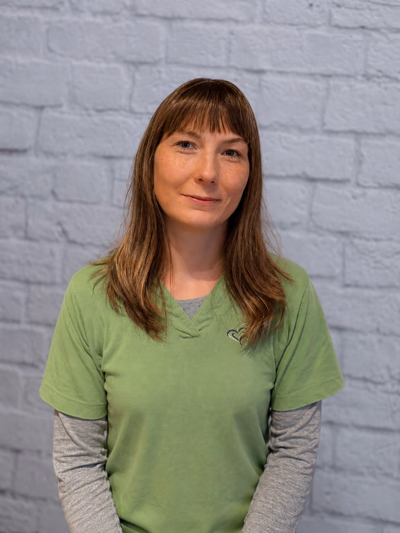 Woman with brown hair wearing a green shirt and gray long sleeves, smiling slightly against a gray brick wall.