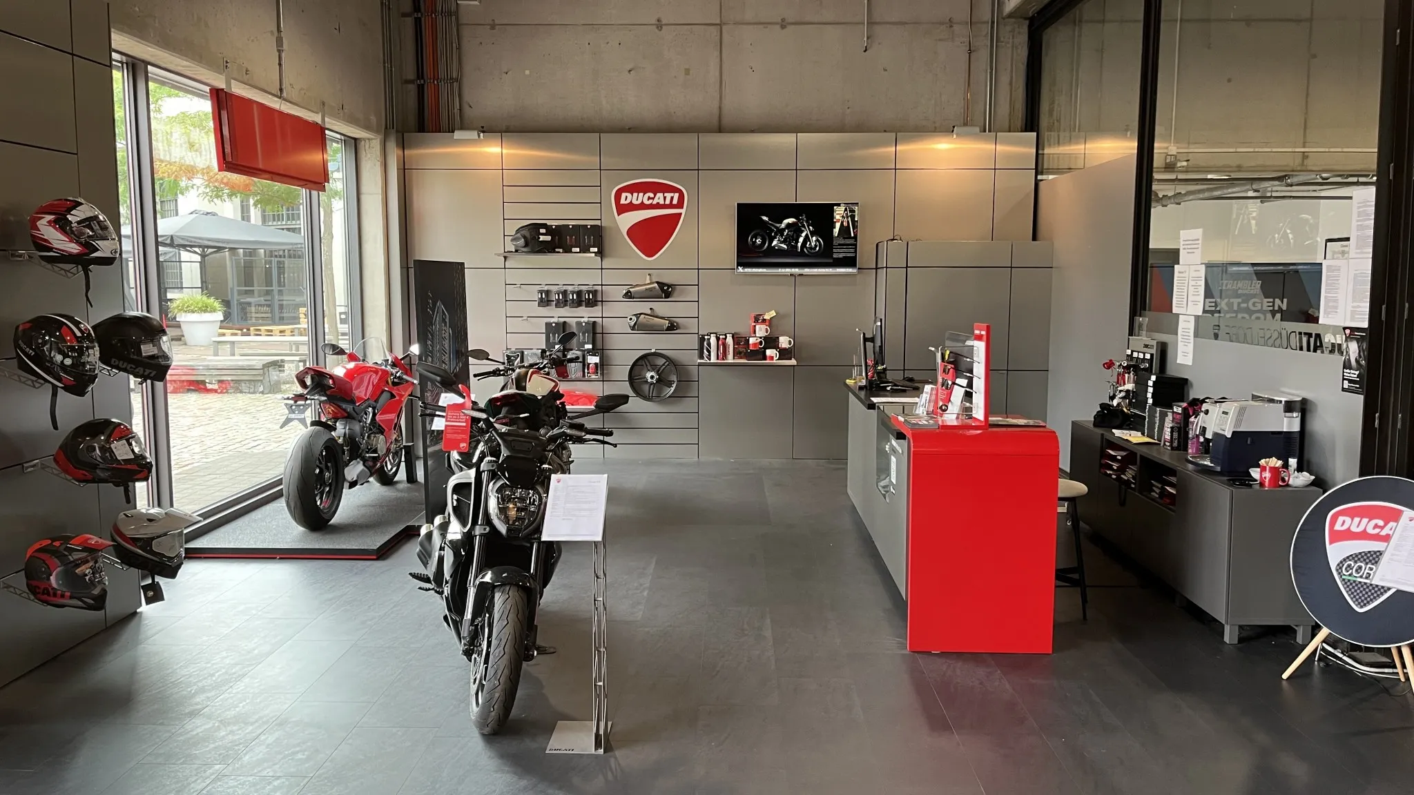 Innenansicht eines Ducati Stores mit ausgestellten Motorrädern, Helmen und Zubehör. Vorne eine schwarze Ducati, hinten eine rote Ducati, daneben ein Verkaufs- und Beratungsbereich.