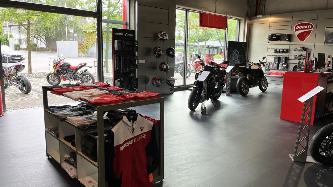 Innenansicht eines Ducati Stores mit ausgestellten Motorrädern, Ducati Corse Bekleidung und Helmen. Vorne ein Verkaufstisch mit Merchandising, im Hintergrund schwarze und rote Ducati Motorräder.