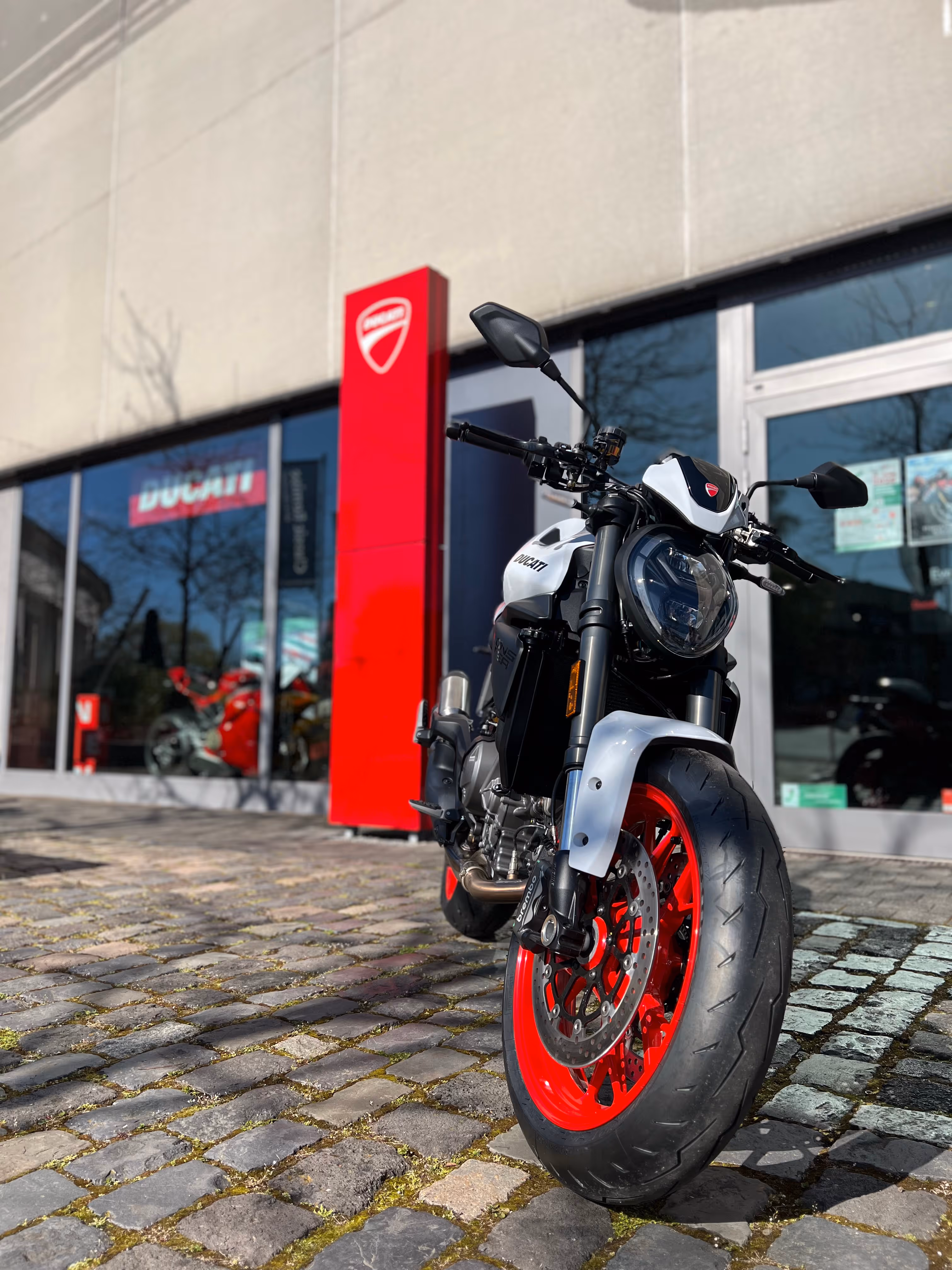 Weißes Ducati-Motorrad mit roten Felgen vor einem Ducati-Händler mit Glasfassade und rotem Schild.