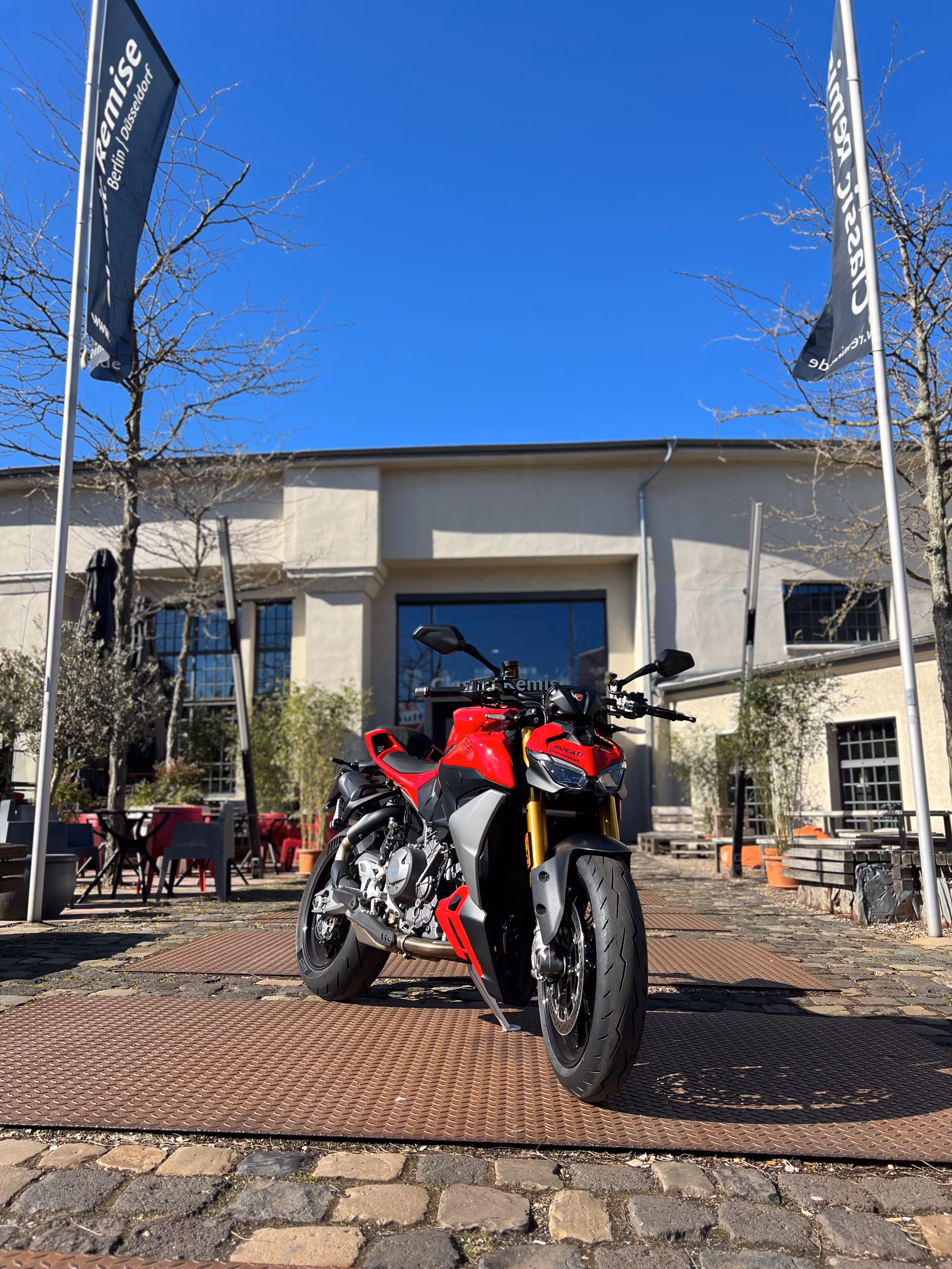 Rotes Ducati-Motorrad auf gepflastertem Boden vor einem hellen Gebäude mit blauen Himmel.