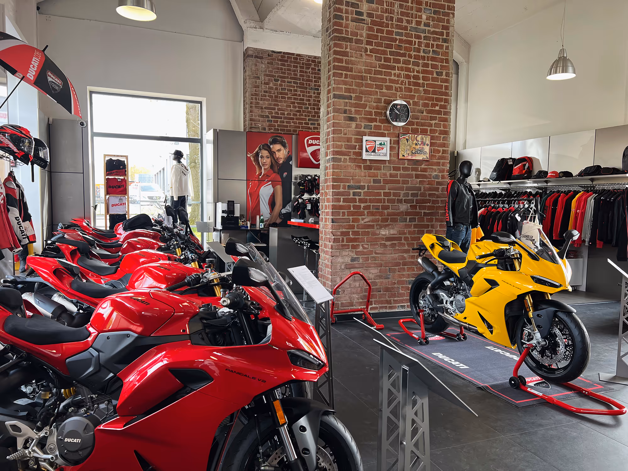Innenraum eines Ducati-Motorradgeschäfts mit mehreren roten Motorrädern in einer Reihe und einem gelben Motorrad auf einem Ständer, umgeben von Ducati-Bekleidung und Zubehör.