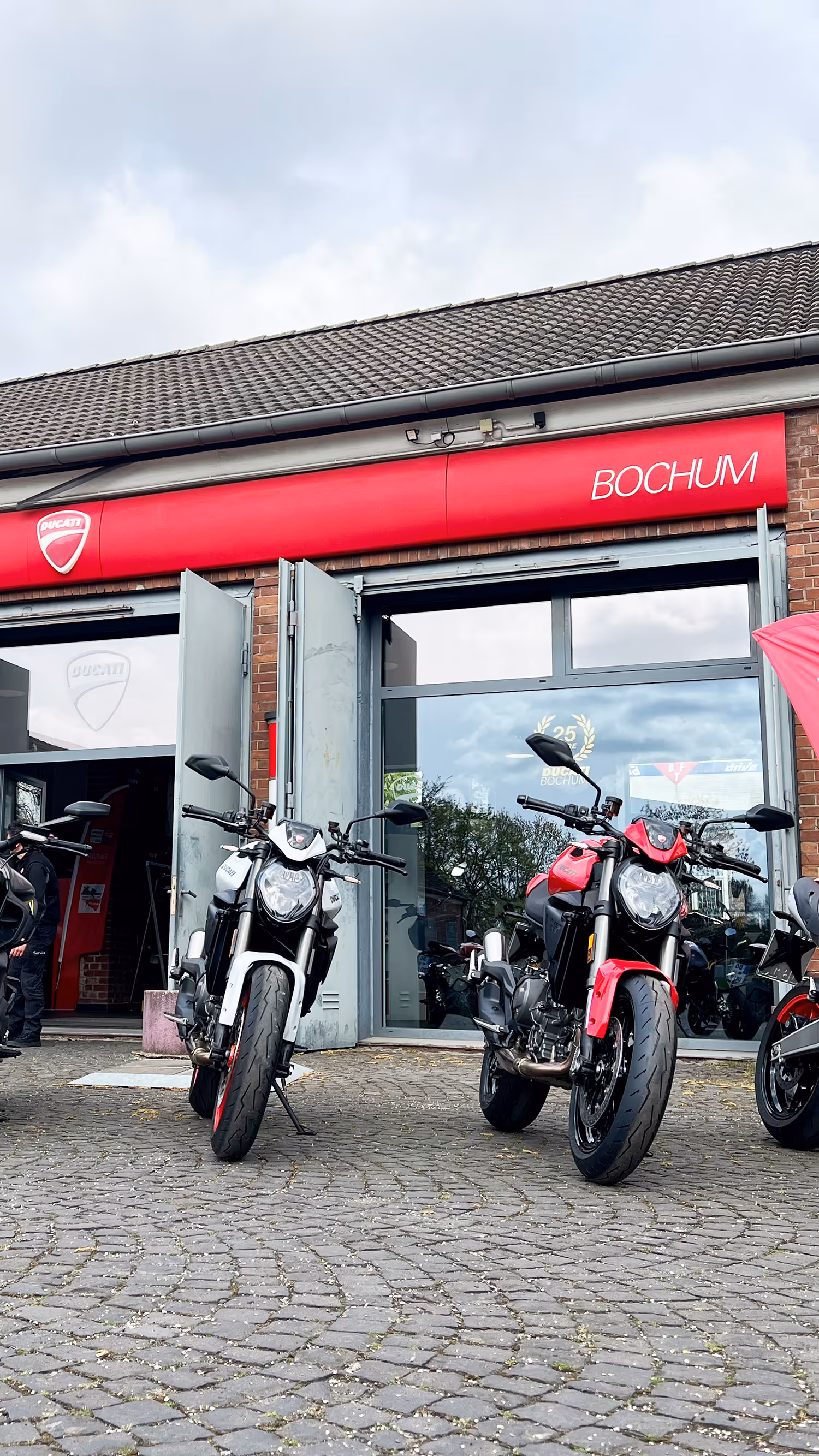 Weißes Ducati-Motorrad mit roten Felgen vor einem Ducati-Händler mit Glasfassade und rotem Schild.