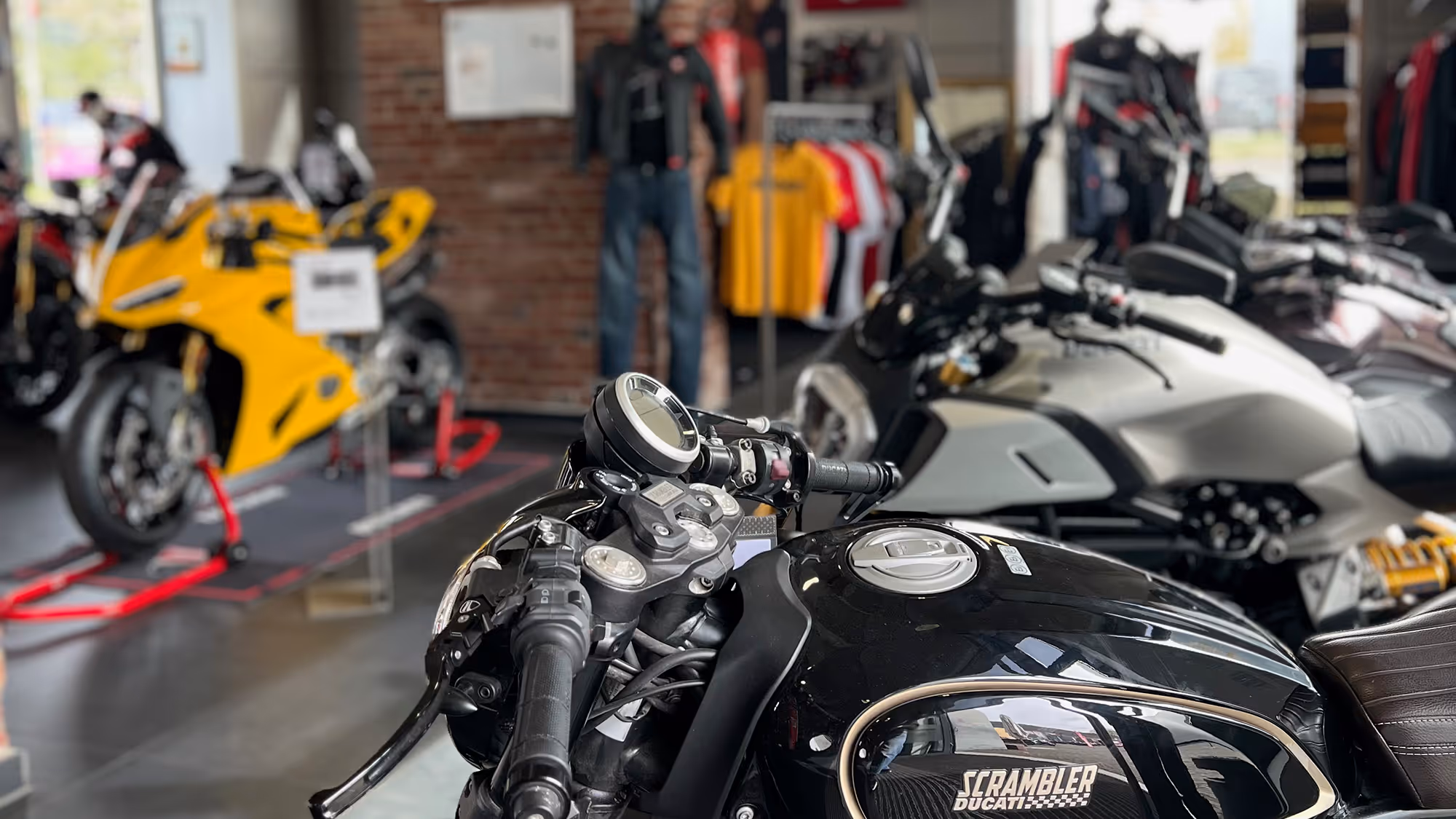 Nahaufnahme eines schwarzen Ducati Scrambler Motorrads in einem Motorradgeschäft mit anderen Motorrädern und Bekleidung im Hintergrund.