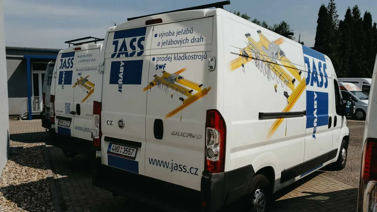 Bílé dodávky s reklamou JASS jeřáby a obrázkem žlutého jeřábu zaparkované vedle sebe.