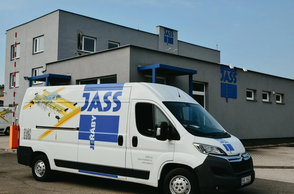 Bílý dodávkový automobil s logem JASS parkuje před šedou budovou firmy JASS.