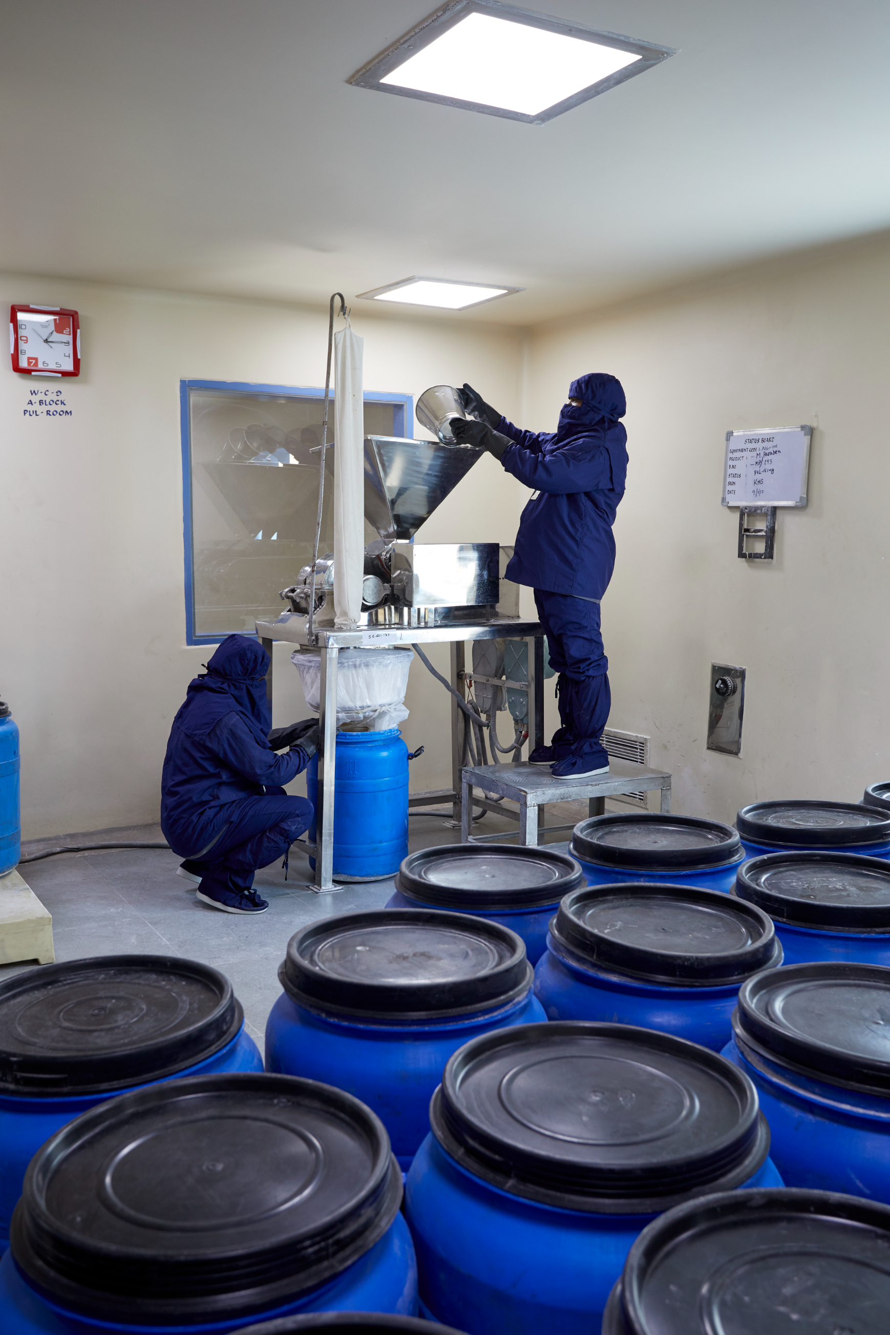 Operators filling chemical products into drums using enclosed processing equipment in a controlled manufacturing environment.