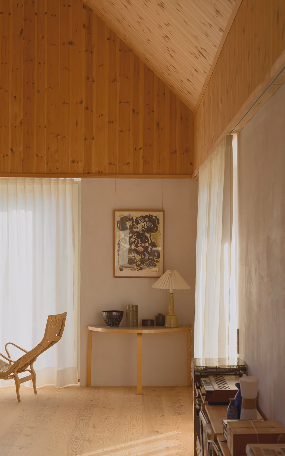 Et lyst, minimalistisk rum med trægulv og vægpaneler. Et smalt, halvcirkelformet bord står mod en væg med en abstrakt rammet maleri hængende over. På bordet står en beige bordlampe med foldet skærm og flere dekorative krukker. To store vinduer med tynde, hvide gardiner slipper blødt lys ind. En fritstående træstol med flettet sikkerhed er til venstre. En reol med bøger og en vintage grammofon er til højre.