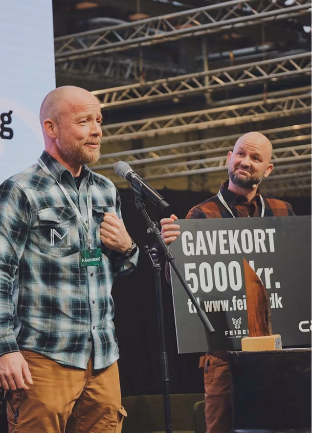 To mænd står på en scene med metalkonstruktioner i loftet. Den ene mand taler i en mikrofon og har på en ternet skjorte og brune bukser. Den anden mand, også i ternet skjorte, holder et stort gavekort med teksten "GAVEKORT 5000 kr." og et websted, begge smilende.
