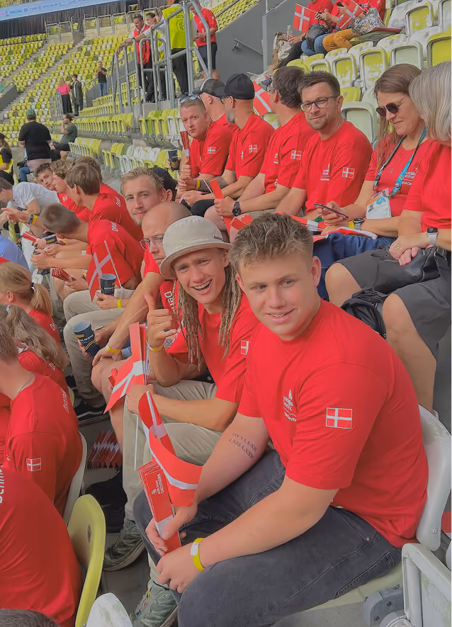 En gruppe mennesker i røde T-shirts med Danmarks flag sidder på gul-grønne stadionpladser, nogle med danske flag i hænderne, mens de smiler og nyder begivenheden.
