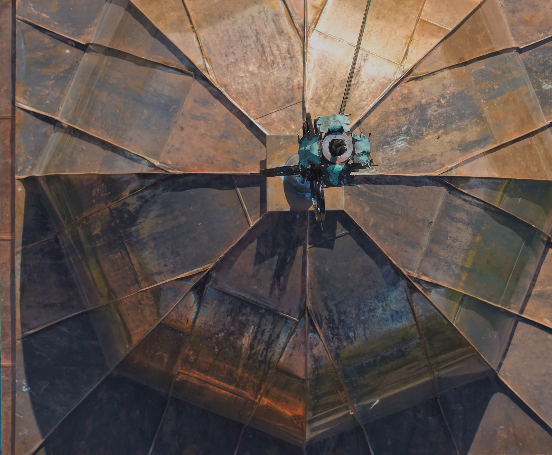 Top-down view of a large, metallic, weathered industrial structure with a central hub and radiating panels casting sharp shadows.
