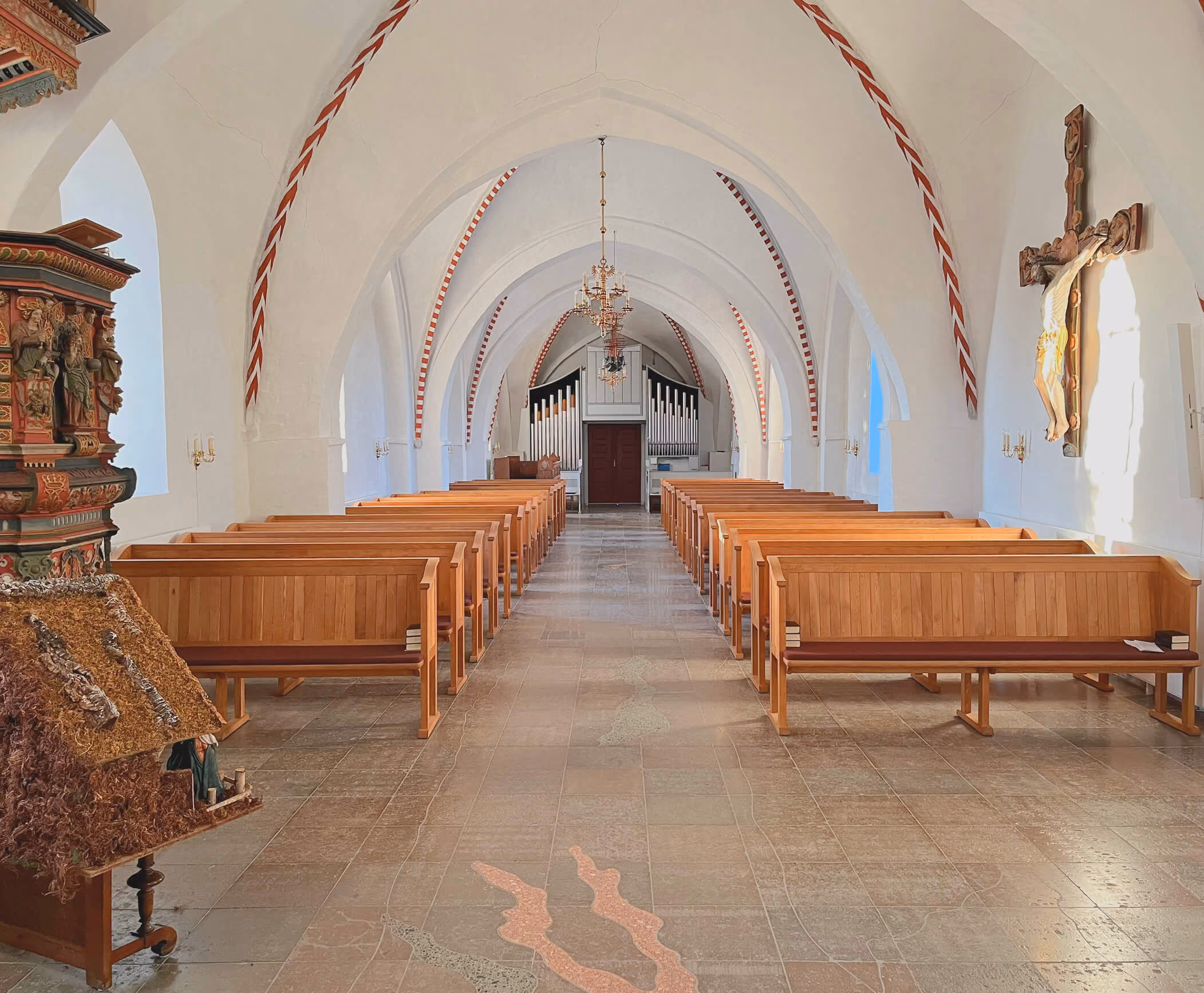 Indvendigt billede af en kirke med hvide hvælvede lofter, røde dekorative striber langs buerne, rækker af træbænke på hver side af midtergangen, et orgel i bagenden og et stort trækrucifiks på højre væg.