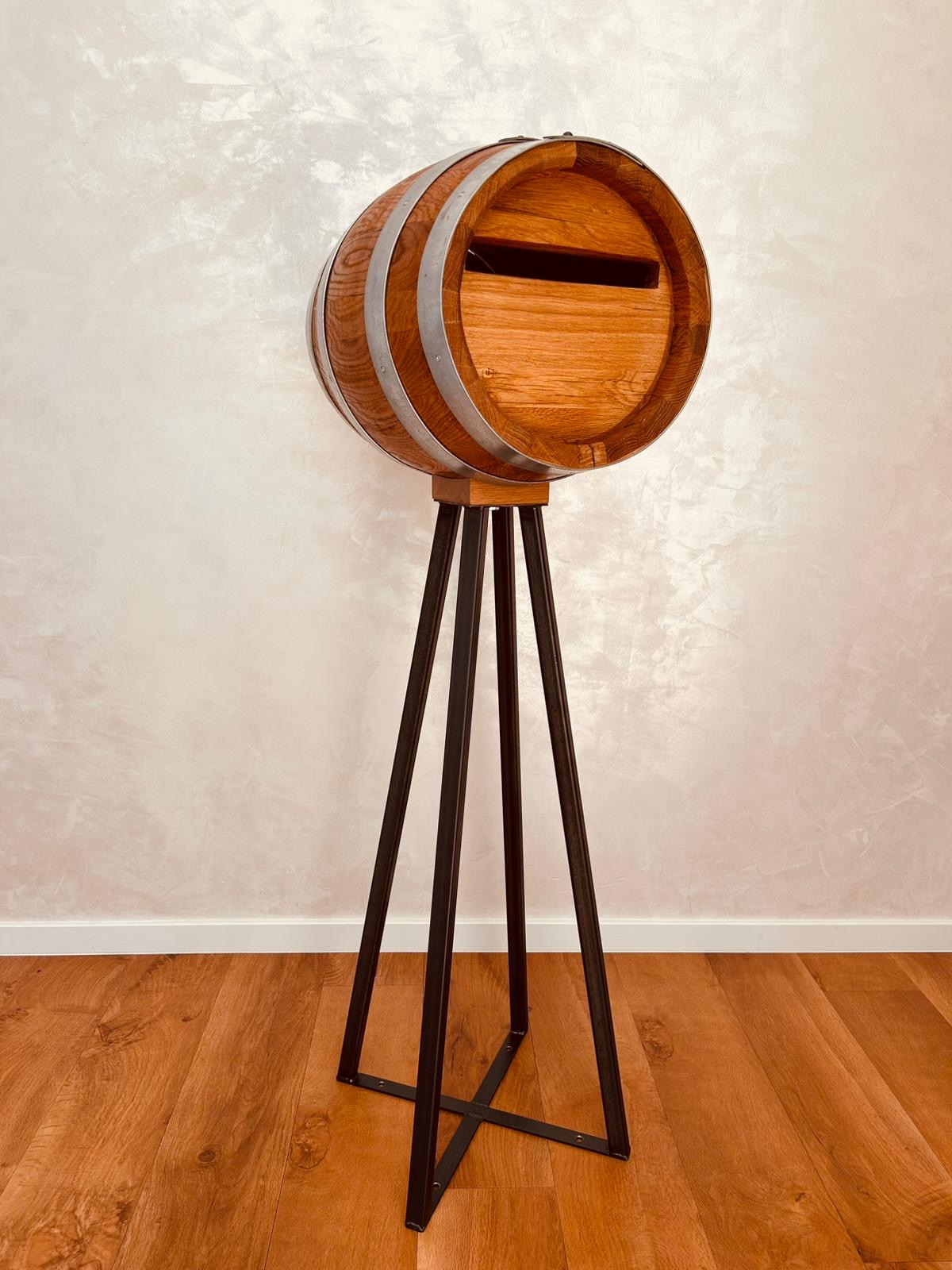 Wooden barrel with metal bands mounted on black metal tripod stand against beige wall.