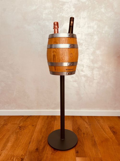 Wooden barrel wine cooler on black metal stand with two bottles inside on wooden floor.