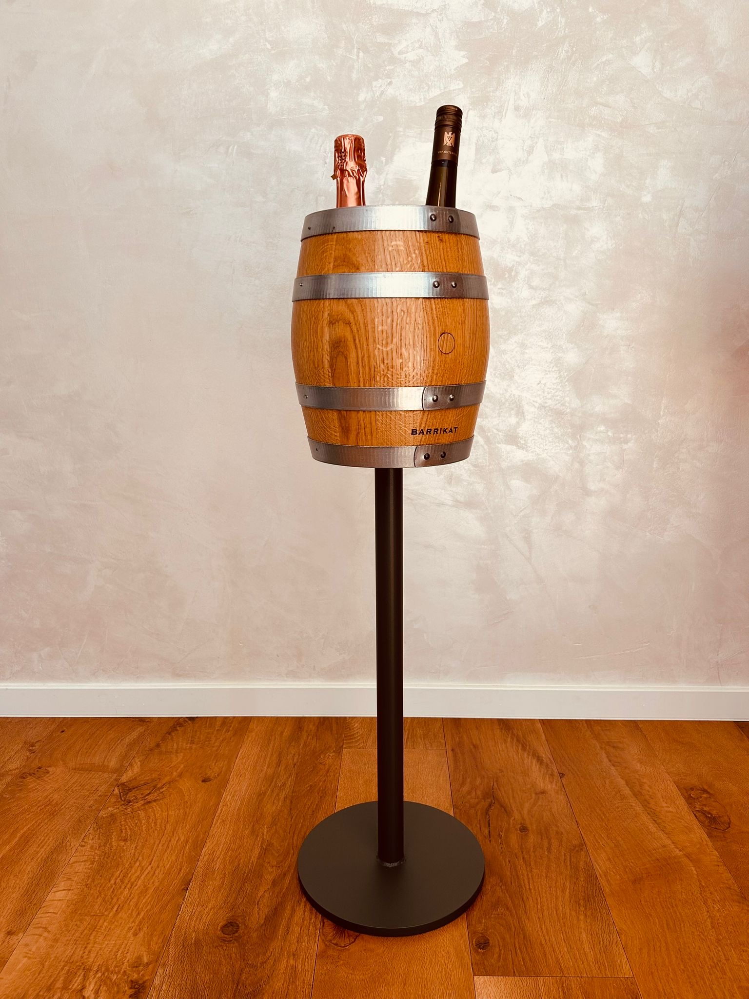 Wooden barrel wine cooler on black metal stand with two bottles inside on wooden floor.