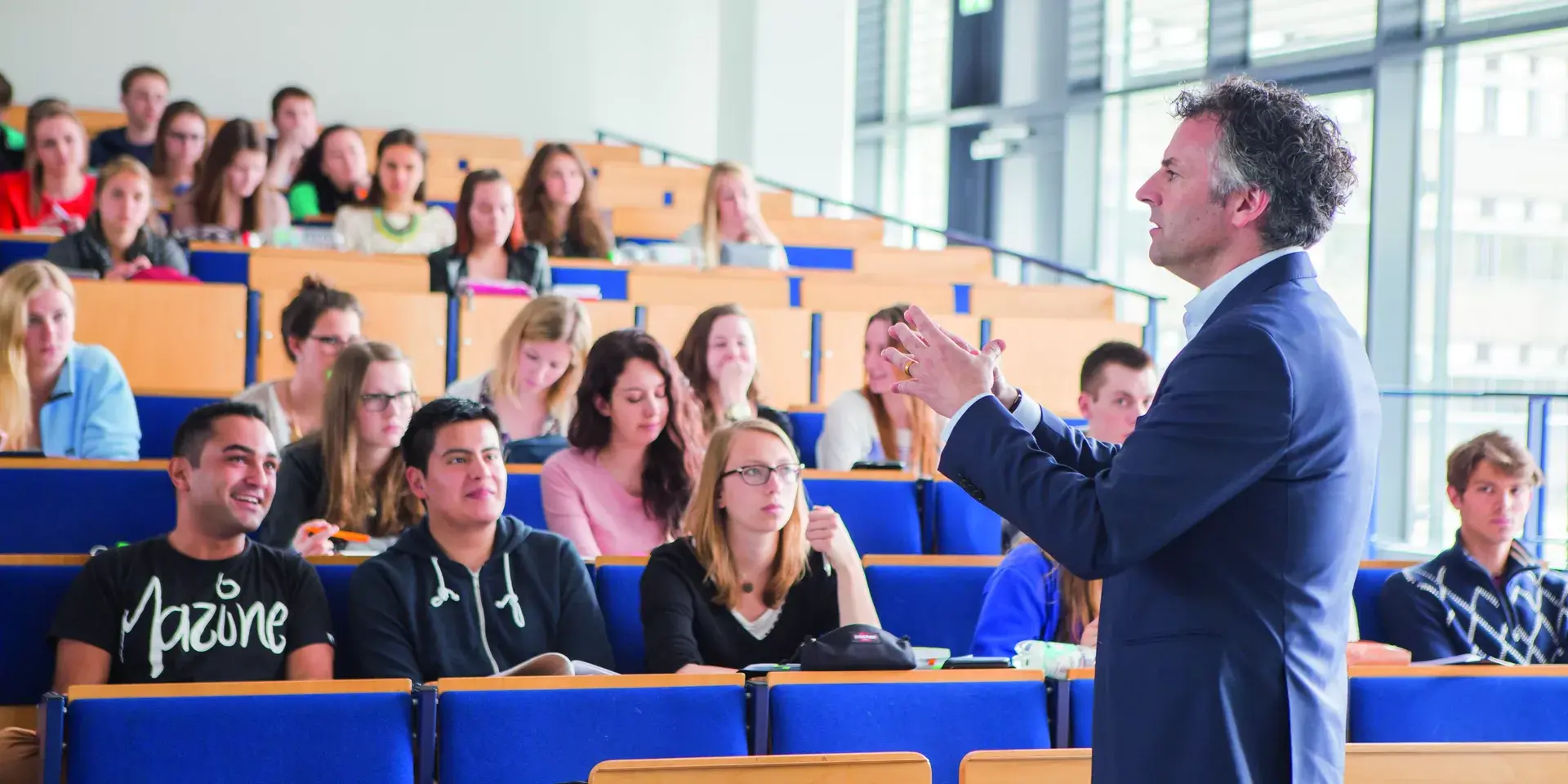 Male professor in a suit speaking to attentive students seated in a tiered university lecture hall.