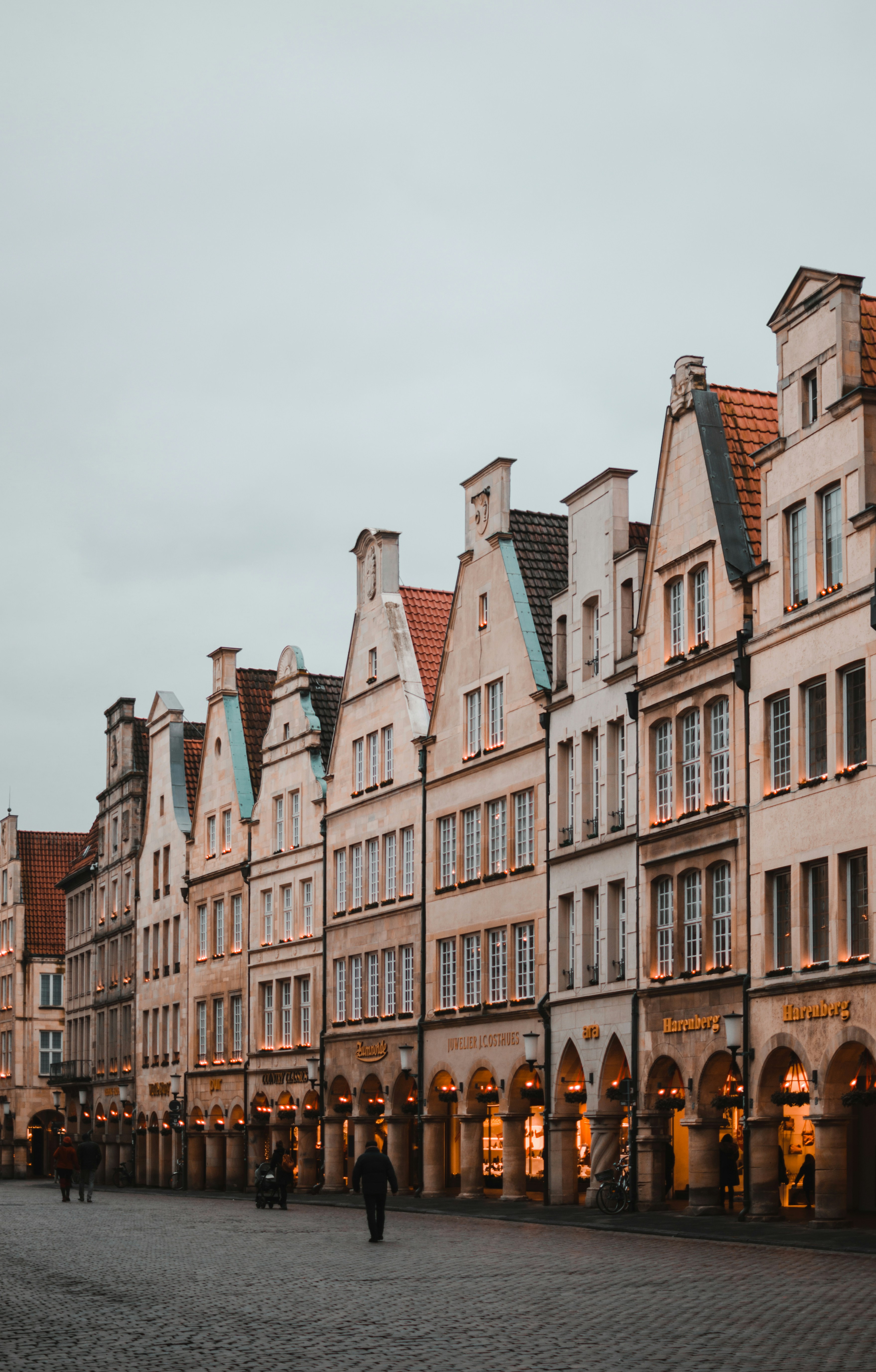 Row of historic gabled buildings with lit storefronts and cobblestone street under gray sky.